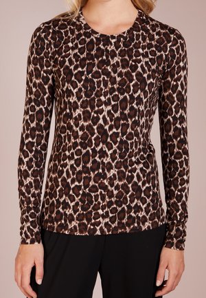 Frau trägt ein eng anliegendes Langarm-Top mit Leopardenmuster und rundem Ausschnitt sowie schwarze Hosen, stehend vor einem neutralen Hintergrund.