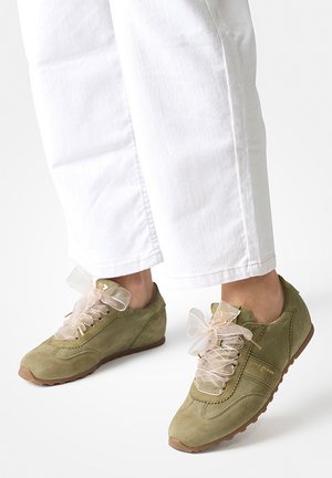 Person trägt weiße Hose und olivgrüne Wildleder-Sneaker mit großen durchsichtigen Schleifenbändern und steht auf einer weißen Oberfläche.