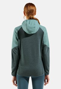 Veste à capuche avec un corps gris foncé et des accents sarcelle, fabriquée à partir d'un tissu doux et texturé, présentant une coupe décontractée et un design athlétique.