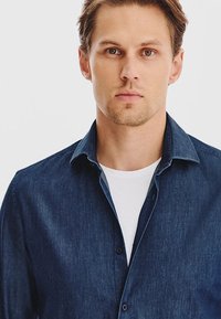 Koszula dżinsowa w ciemnoniebieskim kolorze, z długim rękawem, spiczastym kołnierzykiem, zapinana na guziki, o miękkiej fakturze. Nosi się ją na białej koszulce.