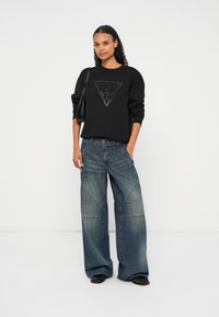 Černá mikina s trojúhelníkovým logem, doplněná širokými džínami s tónovým prošíváním a vybledlou modrou barvou. Černé boty.