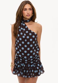 Femme portant une robe noire à une épaule avec de grands pois bleus et un ourlet volanté, accessoirisée avec des bracelets et des boucles d’oreilles en or.