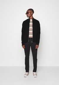 Chaqueta negra con cremallera sobre una camisa de rayas, pantalones oscuros y zapatillas color beige. Modelo usando gafas de sol, de pie contra un fondo blanco.