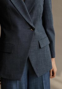 Detailní záběr na osobu ve tmavě šedém blejzru na jedno knoflík s klopovými kapsami a odpovídajících modrých kalhotách.