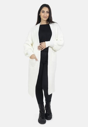 Cardigan lungo bianco a maglia con design aperto, texture a coste e due tasche frontali, abbinato a una maglietta e pantaloni neri e stivali neri.