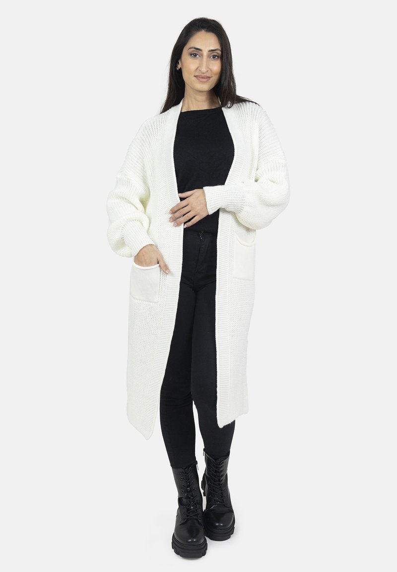 Long gilet en tricot blanc avec un design à ouverture frontale, texture côtelée, et deux poches avant, associé à un haut noir et à un pantalon noir, ainsi qu'à des bottes noires.