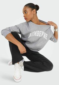 Mujer sentada en el suelo mirando de lado, vestida con una sudadera gris con el texto "NUMBERNE", pantalones negros, zapatillas blancas, pendientes de aro y pulseras.