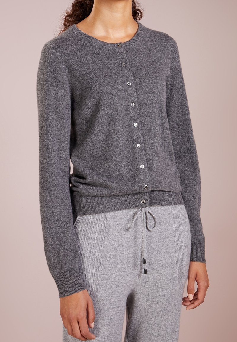 Cardigan en laine grise avec un col rond, une fermeture à boutons sur le devant et des poignets côtelés. Associé à un pantalon gris à cordon de serrage avec un motif côtelé vertical.