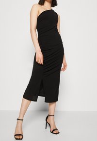 Kobieta w czarnej, bezrękawnikowej sukience midi z bocznym rozcięciem, stojąca w czarnych sandałach na wysokim obcasie na jednolitym białym tle.