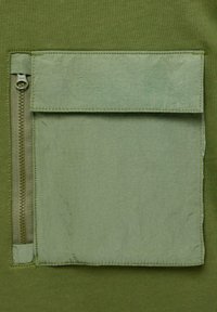 Olivgrünes Oberteil mit einer strukturierten Taschenklappe mit Reißverschluss, ergänzt durch einen glatten Stoff und gestickte Akzente.