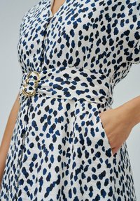 Bílá látková šaty s modro-černým leopardím potiskem, krátké rukávy, knoflíková přední část a opasek v pase s zlatou sponou. Obsahuje kapsy.