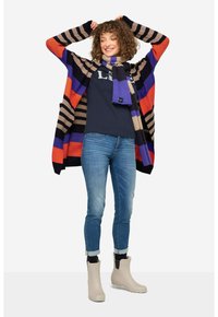 Färgglad randig kofta i röd, lila och beige nyanser, kombinerad med en marinblå topp, blå jeans och beige regnstövlar.