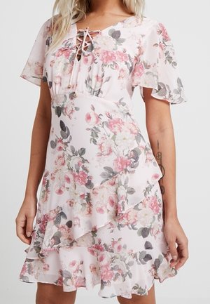 Kobieta ubrana w krótką, różową kwiecistą sukienkę kopertową z falbaniastymi rękawami i dekoltem z wiązaniem, stojąca na jednolitym tle.