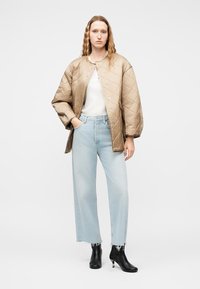 Γυναίκα στέκεται φορώντας μπεζ μπουφάν με καπιτονέ σχέδιο, λευκό τοπ, ανοιχτό μπλε τζιν παντελόνι φαρδιάς γραμμής και μαύρες μποτάκια με τακούνι.