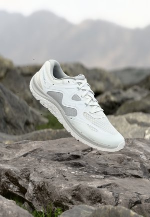 Zapatilla de trail running blanca de Merrell suspendida sobre terreno rocoso con un fondo montañoso desenfocado.