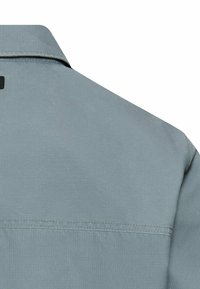 Jasnoszara koszula prezentująca gładką, fakturowaną tkaninę z ostrym kołnierzykiem i widocznym szwem na ramieniu.