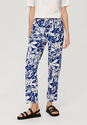Personne portant un pantalon bleu et blanc à imprimé feuille abstrait, avec une taille nouée et des sandales à brides noires, debout devant un fond uni.