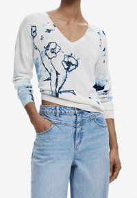 Pull blanc avec un motif floral bleu, col en V et manches longues. Associé à un jean taille haute bleu clair avec des poches latérales.