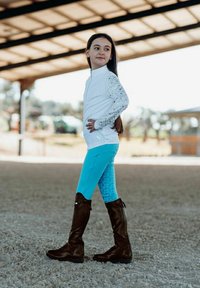 Conjunto ecuestre que incluye una blusa blanca de manga larga con mangas texturizadas, leggings turquesas con un patrón y botas de montar marrones.