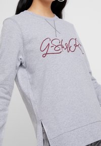Grå sweatshirt med röd broderad "G-Star" logotyp, rundad halsringning, sidofickor och strukturerad tyg. Ribbad detalj vid ärmarna och nederkanten.