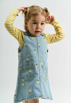 Džínová šaty s vyšívaným žlutým ananasem, zapínání na knoflíky a předními kapsami, nošené přes žluté a bílé pruhované tričko s dlouhým rukávem.