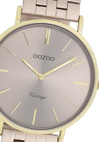 Montre ronde avec un boîtier en métal doré clair et un cadran gris texturé. Comprend des index et des aiguilles dorés, avec un bracelet en métal beige.