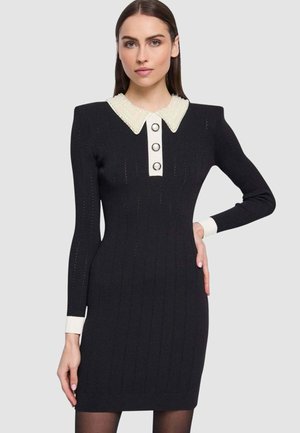 Donna che indossa un vestito aderente in maglia nera con colletto decorato con perle bianche, polsini bianchi e tre bottoni decorativi sul davanti.