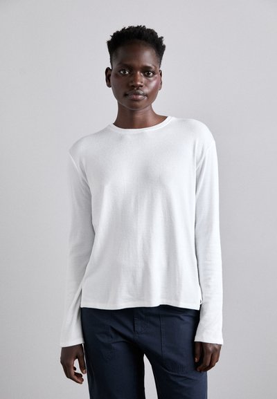 Haut blanc à manches longues en matériau léger, avec un col rond et une coupe décontractée. Présente un ourlet droit et des fentes latérales, porté avec un pantalon sombre.