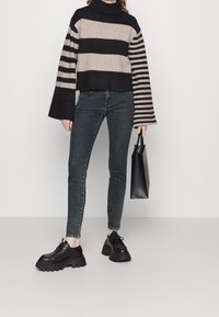 Stickad randig tröja i beige och svart med hög krage. Kombinerad med mörka skinny jeans och svarta klumpiga skor. Håller en svart tote-väska.