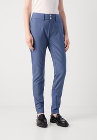 Frau in schlanken blauen Hosen mit doppelter Knopfleiste und schwarzen Slippern, die vor einem schlichten hellen Hintergrund steht.