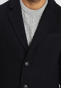 Cappotto in lana blu navy con rever a punta e quattro bottoni, indossato sopra un maglione grigio chiaro in maglia a trecce. Vista ravvicinata del colletto e dei rever.