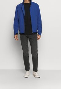 Hombre vestido con chaqueta azul con cremallera, camiseta negra, jeans gris oscuro y zapatillas altas blancas, de pie contra un fondo plano.