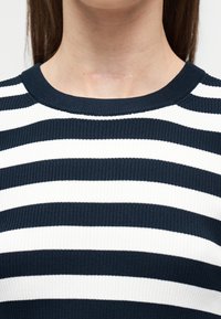 Ουλή στο μπροστινό μέρος του λαιμού ενός ατόμου που φοράει μπλε μπλούζα με ριγέ σχέδιο σε λευκό και μπλε και μακριά καστανά μαλλιά ορατά.