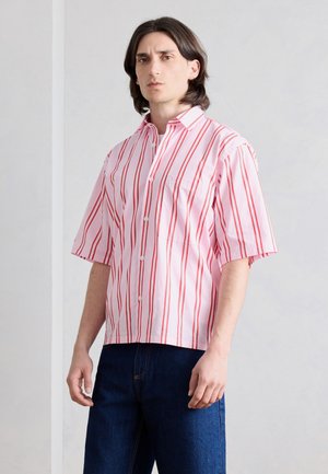 Young man with shoulder-length dark hair wearing a pink short-sleeve button-up shirt with vertical red stripes and dark blue jeans.