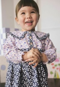 Usmievajúce sa malé dieťa s krátkymi vlasmi, oblečené v kvetinových šatách, spája ruky pred jasným, hravým pozadím.