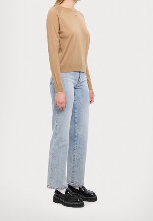 Frau trägt einen langärmeligen Pullover in Kamelhaarfarbe mit kleinem rosa Herz, hellblaue gerade Jeans, weiße Socken und schwarze Loafers, stehend.