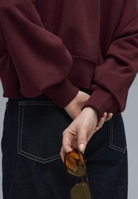 Bordeaux sweatshirt med voluminøse ærmer, parret med mørk denim. Hånd der holder skildpadde-sunglasses med gule linser bag ryggen.