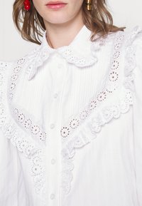 Blusa bianca in tessuto testurizzato, caratterizzata da un ampio colletto con ruches ricamate e bottoni sul davanti.
