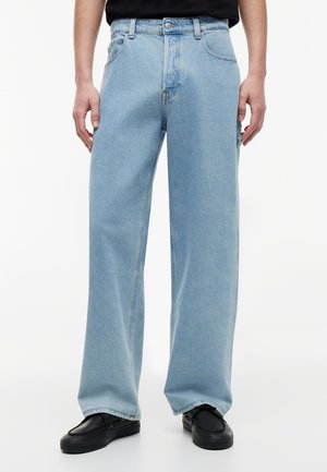 Φαρδιά τζιν σε ανοιχτό γαλάζιο χρώμα, φορεμένα με μαύρα παπούτσια χωρίς κορδόνια, μοντέλο όρθιο με τα χέρια χαλαρά δίπλα στο σώμα σε λευκό φόντο.