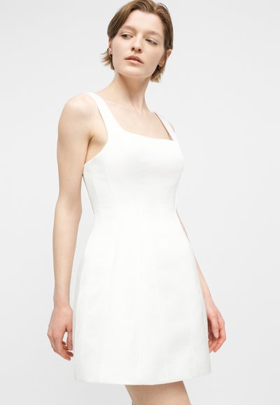 Jeune femme portant une robe blanche sans manches, ajustée, longueur genou, debout devant un fond clair uni.
