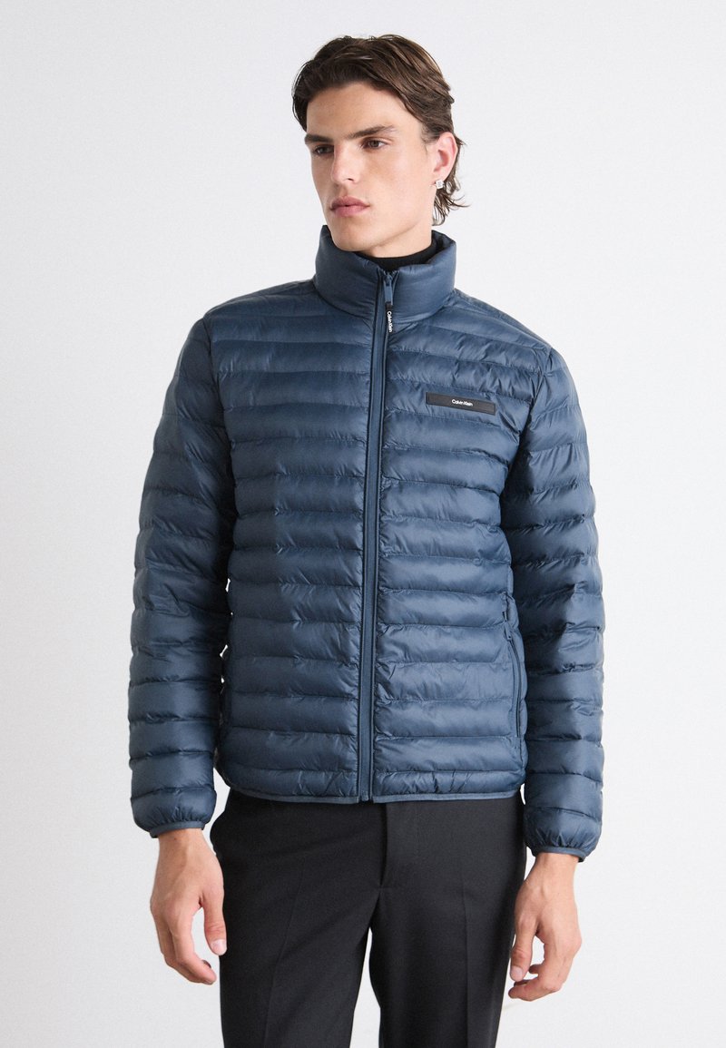 Giacca a vento blu navy con zip frontale, colletto alto e motivo trapuntato orizzontale. Presenta un piccolo logo sul petto.