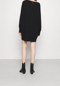Robe-pull noire oversize avec une coupe ample, des manches longues, et un détail discret de logo doré au dos. Associée à des bottines noires.