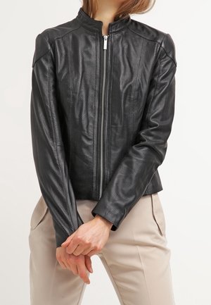 Musta nahkatakki, jossa on eteen vetoketju ja pienet reiät, yllään beigehousut, kädet ristissä vyötäröllä.