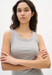 Hallitusseente tekstuuri ja laia kaelusega hall sobiv kattepealne, mis on ilma varrukateta. Mudel poseerib käed ristis valge tausta ees.