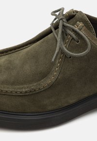Zapato de ante verde oliva con punta redondeada, superficie texturizada, costuras beige y detalles de cordones a juego; cuenta con una suela de goma negra.