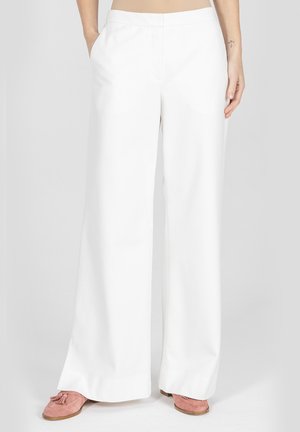 Kobieta ubrana w białe spodnie z wysokim stanem i szerokimi nogawkami z bocznymi kieszeniami oraz różowe mokasyny zamszowe, stojąca na jednolitym jasnym tle.