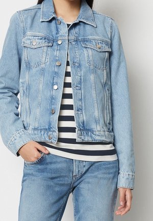 Femme portant une veste en jean bleu clair sur une chemise rayée noir et blanc avec un jean bleu clair assorti, main dans la poche.