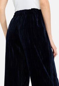 Pantaloni di velluto blu scuro con una texture arricciata, dotati di una vita elastica e gambe larghi, visti da dietro.