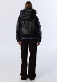 Persoon met lang haar, gekleed in een zwarte gewatteerde jas met capuchon en donkerbruine wijde broek, staand voor een effen lichte achtergrond.