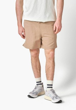 Bărbat care poartă pantaloni scurți bej, tricou alb, șosete albe cu dungi negre și adidași New Balance gri, stând pe un fundal alb.